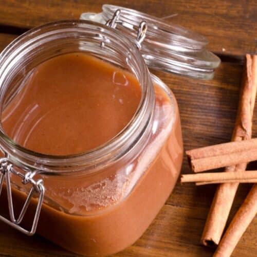 Slow-Cooked Paleo Apple Butter in a glass jar next to cinnamon sticks on a table.